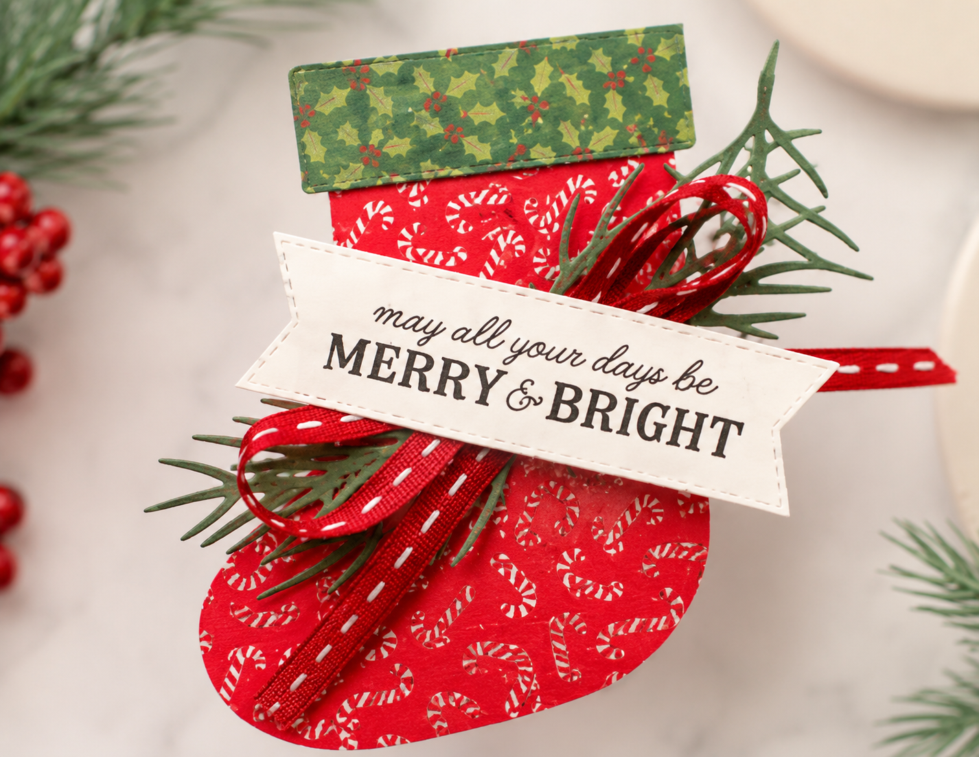 Close-up of a handmade Christmas stocking card embellishment with a Merry & Bright sentiment, red ribbon, and pine branches.