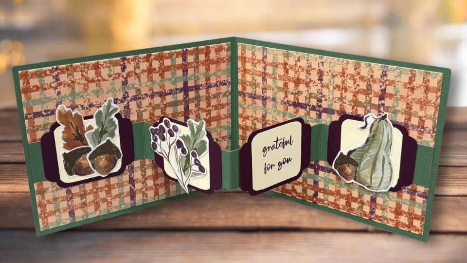 W-Fold card in autumn colors, opened to show four panels with a green plaid background, layered acorn and gourd die cuts, a purple floral accent, and a 'grateful for you' sentiment panel, displayed on a wood surface