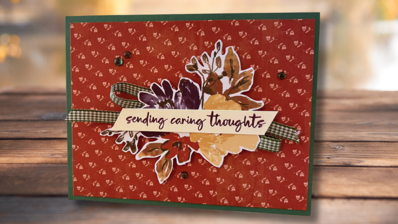 Fall-themed W-fold card with a red floral patterned background and green border, layered leaf and floral die cuts in purple, gold, and rust tones, a gingham ribbon, gold sequins, and a 'sending caring thoughts' sentiment banner, displayed on a wood surface